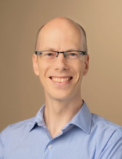 Smiling person with glasses, wearing a blue striped shirt, against a beige background.