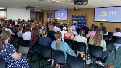 Audience seated in a conference room listening to a speaker at a podium, with two projection screens.