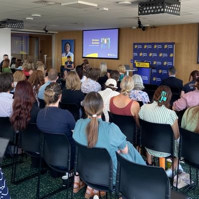 Audience seated in a conference room listening to a speaker at a podium, with two projection screens.