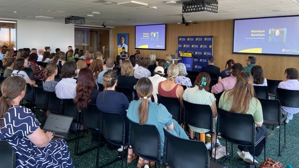 Audience seated in a conference room listening to a speaker at a podium, with two projection screens.