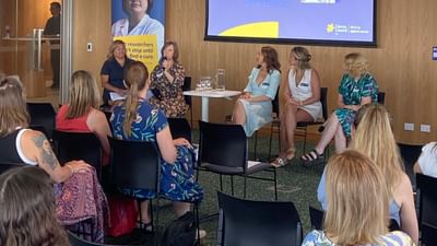Panel of five people on stage in a wood-paneled room; projector screen shows presenter names.