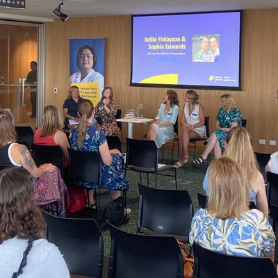 Panel of five people on stage in a wood-paneled room; projector screen shows presenter names.