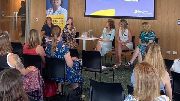 Panel of five people on stage in a wood-paneled room; projector screen shows presenter names.
