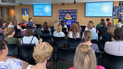 Audience seated in a conference room watching a speaker at a podium with two screens.