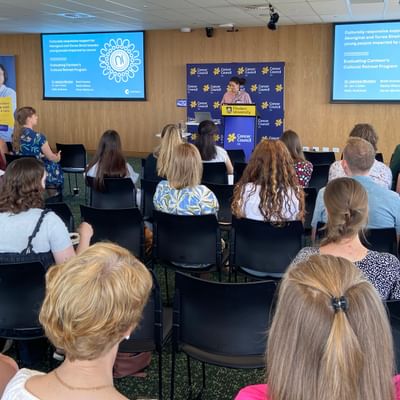 Audience seated in a conference room watching a speaker at a podium with two screens.