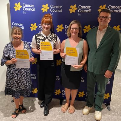 Four people stand in front of a Cancer Council SA backdrop, holding award certificates.