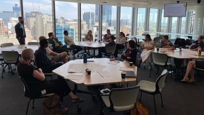 People seated around round tables in a glass-walled conference room overlooking a city skyline.