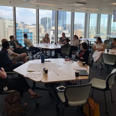 People seated around round tables in a glass-walled conference room overlooking a city skyline.