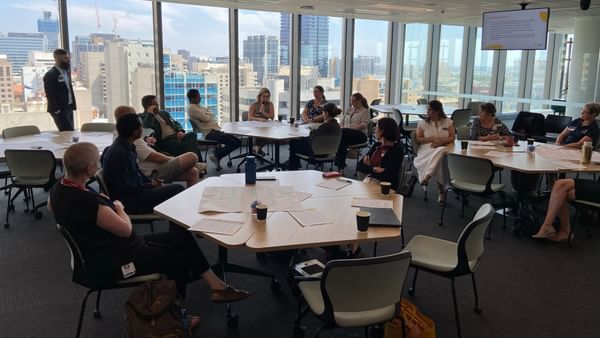 People seated around round tables in a glass-walled conference room overlooking a city skyline.