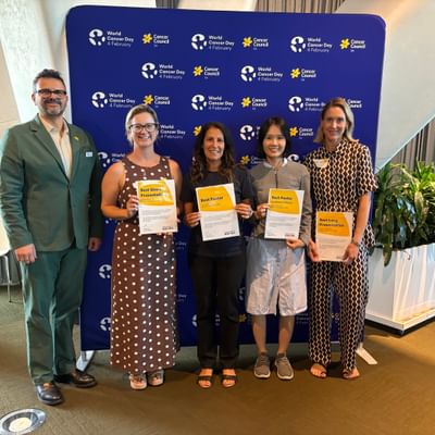 Five people standing in front of a World Cancer Day backdrop, four of them with certificates.