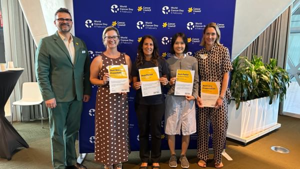 Five people standing in front of a World Cancer Day backdrop, four of them with certificates.