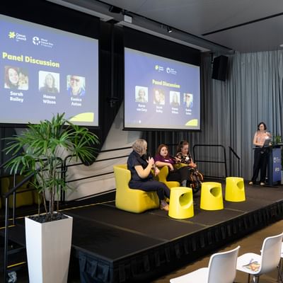 Four people on stage at the Research Symposium. Three of the people are sitting on a discussion panel and one person is standing at a podium asking them questions.