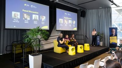 Four people on stage at the Research Symposium. Three of the people are sitting on a discussion panel and one person is standing at a podium asking them questions.
