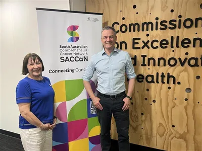 A short woman and a taller man stand either side of a banner depicting the South Australian Comprehensive Cancer Network logo