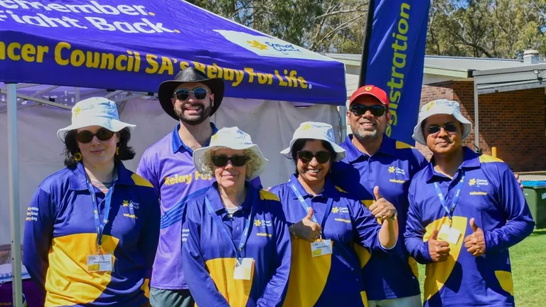 A group of six volunteers, all of different ages and genders, stand in front of a purple Cancer Council SA marquee