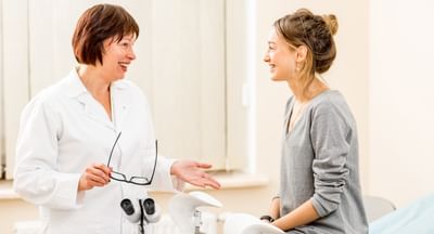 A female doctor on the left speaks to a female patient sitting on the edge of an examination table