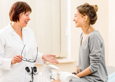 A female doctor on the left speaks to a female patient sitting on the edge of an examination table