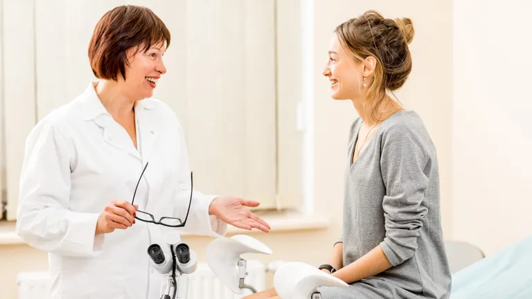 A female doctor on the left speaks to a female patient sitting on the edge of an examination table