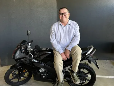 A man wearing glasses sits side-saddle on a motorcycle