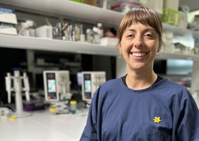 A female cancer researcher smiles at the camera in front of a table covered in laboratory research equipment