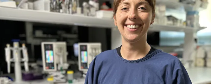 A female cancer researcher smiles at the camera in front of a table covered in laboratory research equipment