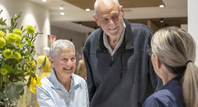 A receptionist, seen from behind, interacts with a short elderly woman and a tall elderly man, both of whom are smiling