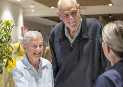 A receptionist, seen from behind, interacts with a short elderly woman and a tall elderly man, both of whom are smiling