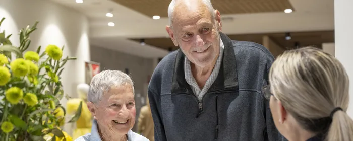 A receptionist, seen from behind, interacts with a short elderly woman and a tall elderly man, both of whom are smiling
