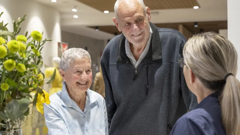 A receptionist, seen from behind, interacts with a short elderly woman and a tall elderly man, both of whom are smiling