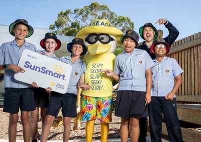 Sid the Seagull stands in the middle of a group of 6 primary school students, with the two students on the left holding a SunSmart sign