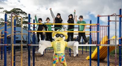 Sid the Seagull stands in front of a school playground, which holds five students from Lake Windemere School