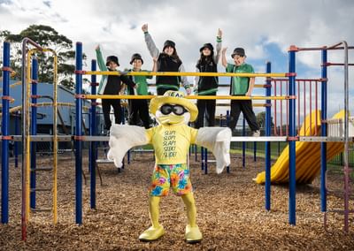 Sid the Seagull stands in front of a school playground, which holds five students from Lake Windemere School