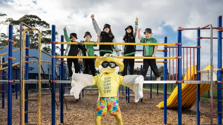 Sid the Seagull stands in front of a school playground, which holds five students from Lake Windemere School