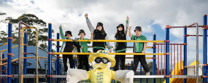 Sid the Seagull stands in front of a school playground, which holds five students from Lake Windemere School
