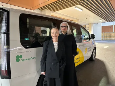 Two women stand in front of the GenesisCare logo located near one of the tail lights of the Cancer Council SA Transport to Treatment van.