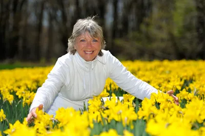 Smiling person in a white jacket reaching through a field of bright yellow daffodils.