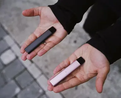 Two hands hold slim rectangular devices, one dark and one pale pink, against a pavement background.