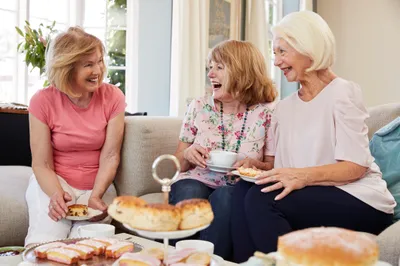 Three seniors sit on a sofa, sharing tea and scones, in a bright living room.