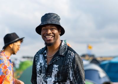 Smiling person in a black patterned shirt and bucket hat at an outdoor event.